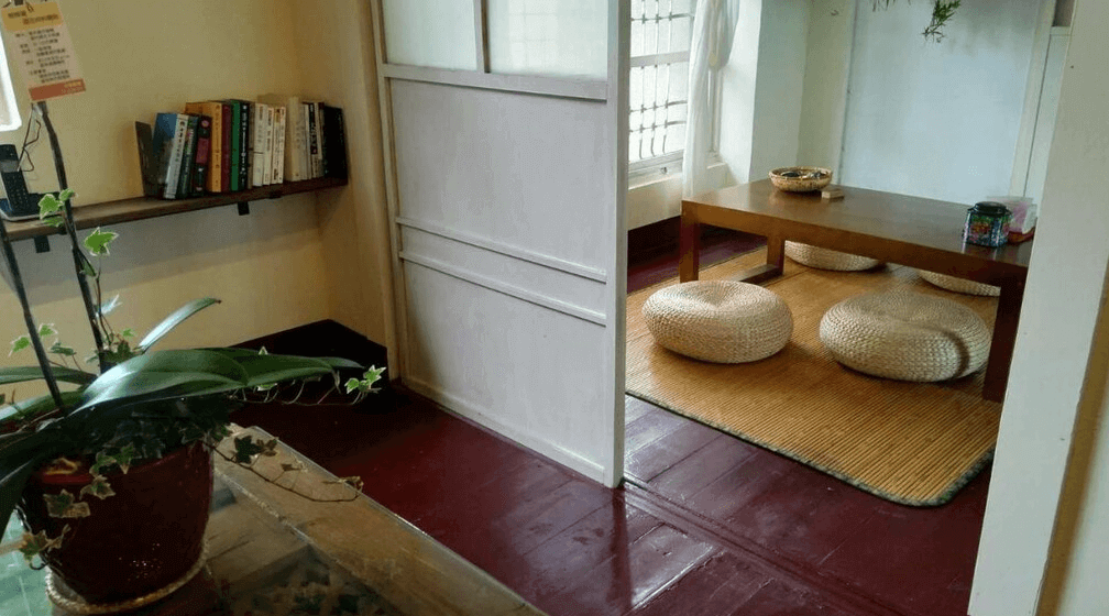 【左營住宿】丈引小院日式榻榻米客廳空間，室內公共區設有榻榻米地墊與木質茶几，搭配藤編坐墊與書架，營造靜謐閱讀與交流氛圍。