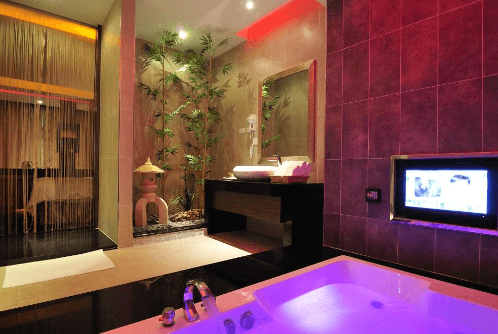 【龍潭住宿】伊加伊時尚汽車旅館的浴室設有大理石按摩浴缸，牆面採紫紅色磁磚與氛圍燈光搭配，呈現浪漫風格。浴缸旁設有電視與乾濕分離設計，洗手台區結合日式植栽造景與燈籠造型擺飾，提升整體質感與放鬆感。