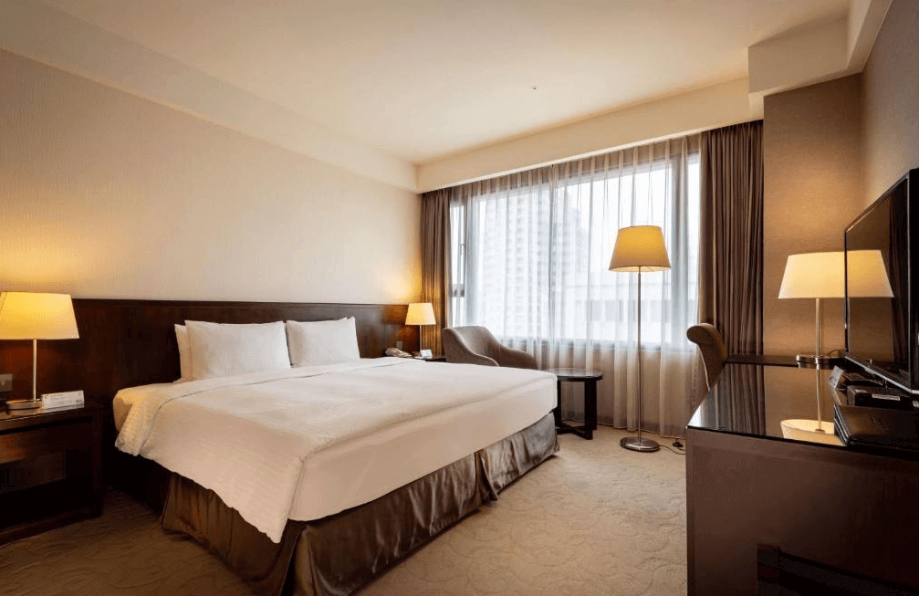 【一中街住宿】來來商旅的雙人房，配有一張加大雙人床與柔和黃光燈飾，大面窗景引入自然光線，房內設有沙發與辦公桌，空間寬敞舒適。