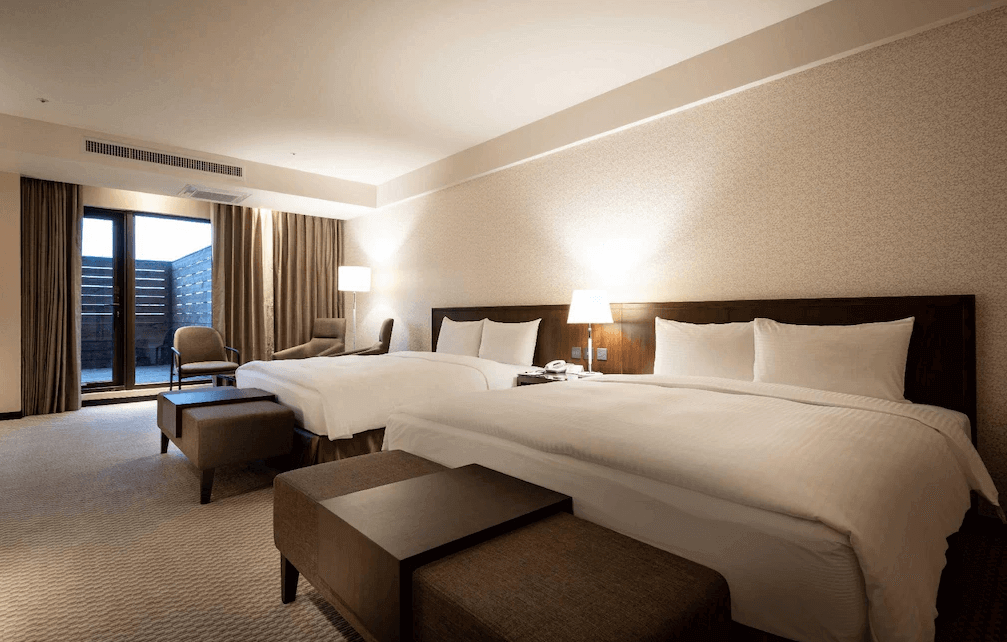 【一中街住宿】來來商旅的四人房，設有兩張雙人床，搭配木質家具與暖色系燈光，空間內配有沙發椅、茶几與大片窗戶，營造溫暖放鬆的住宿氛圍。