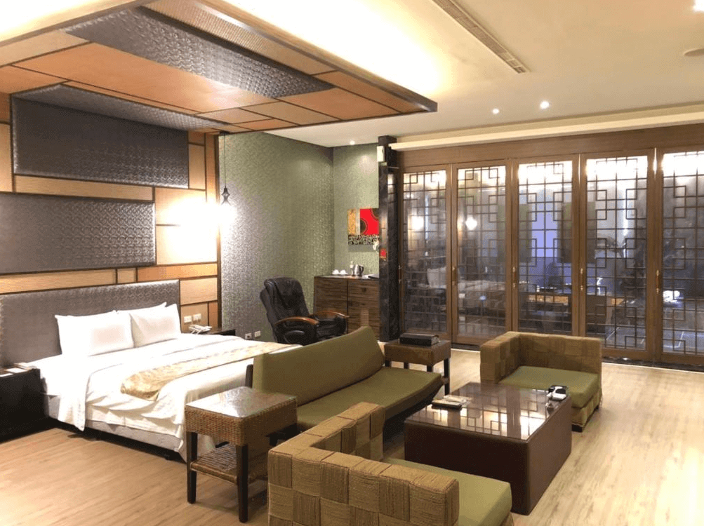 【楊梅住宿】依蝶時尚汽車旅館的套房，設有一張雙人床與多組藤編沙發組，風格融合東方元素與自然材質。床頭牆與天花板設計細緻，大片落地窗配有木質格柵，增添隱私與設計感。