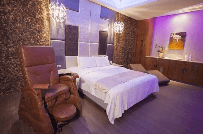 【楊梅住宿】依蝶時尚汽車旅館的房間，配有雙人床、按摩椅與懶人沙發，燈光以紫色與柔黃光營造放鬆氛圍。床頭牆面採用立體材質設計，右側設有木質櫃台與泡茶設備。