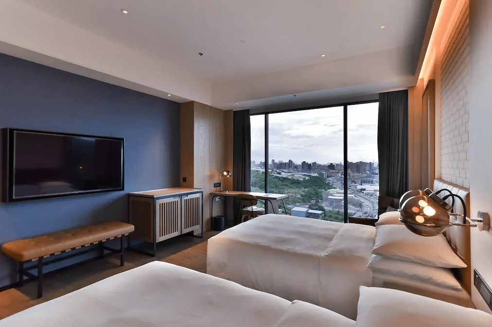 【台北寵物友善住宿】八里福朋喜來登酒店的雙床客房，設有大片落地窗可俯瞰市區景觀，搭配深藍色牆面、皮革長椅、木質家具與書桌，營造沉穩舒適的住宿氛圍。