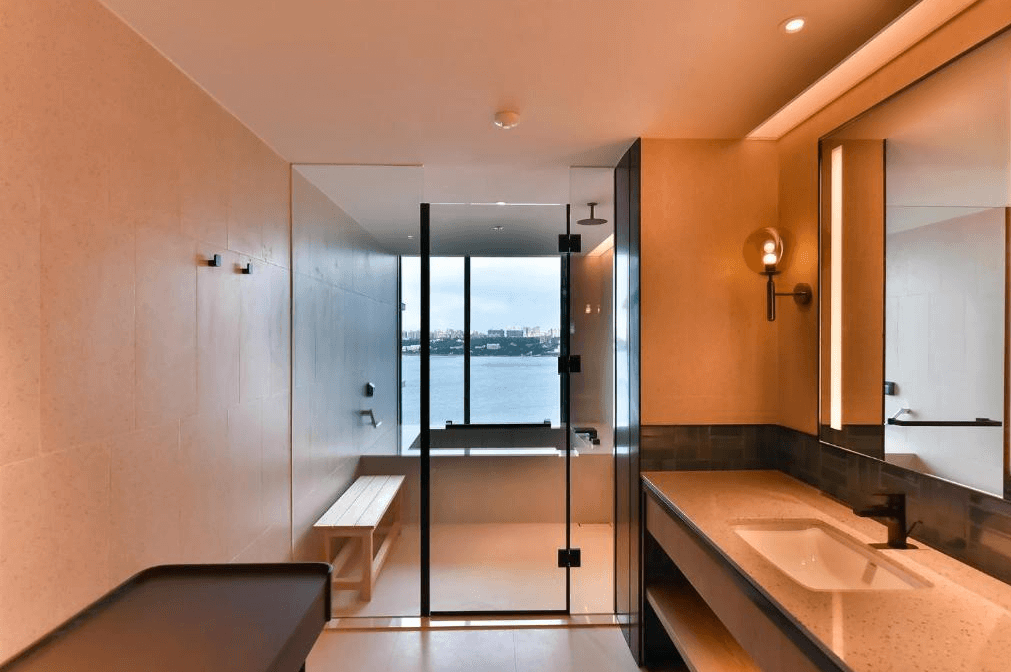 【台北寵物友善住宿】八里福朋喜來登酒店的浴室設計，配有玻璃淋浴間與長型洗手台，大片窗戶可望見水岸景色，搭配木質長凳與暖色照明，展現高級感與放鬆氛圍。