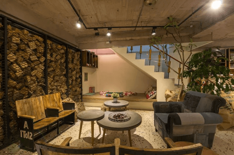 【台北車站青年旅館】北門窩泊旅的交誼廳空間，以原木與工業風結合為主軸，配置木桌椅、沙發及造型燈具，牆面裝飾原木堆疊，一旁還有和式地墊區可供放鬆休息。