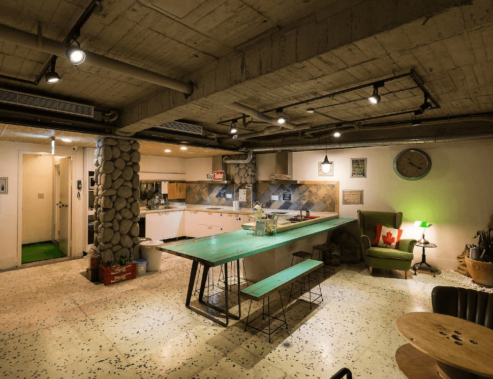 【台北車站青年旅館】北門窩泊旅的開放式廚房空間設有長桌與綠色木質長椅，搭配復古吊燈與裸露水管天花，角落還有舒適沙發與裝飾抱枕，營造出居家感與溫馨氛圍，適合旅客互動用餐。