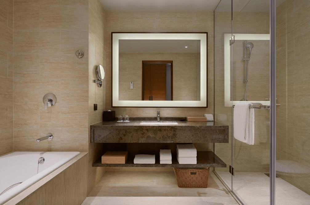 【台北寵物友善住宿】台北六福萬怡酒店的浴室空間，設有獨立浴缸、玻璃淋浴間、大片鏡面與嵌燈，石材洗手台下方整齊擺放毛巾與籃子，整體設計典雅且實用。