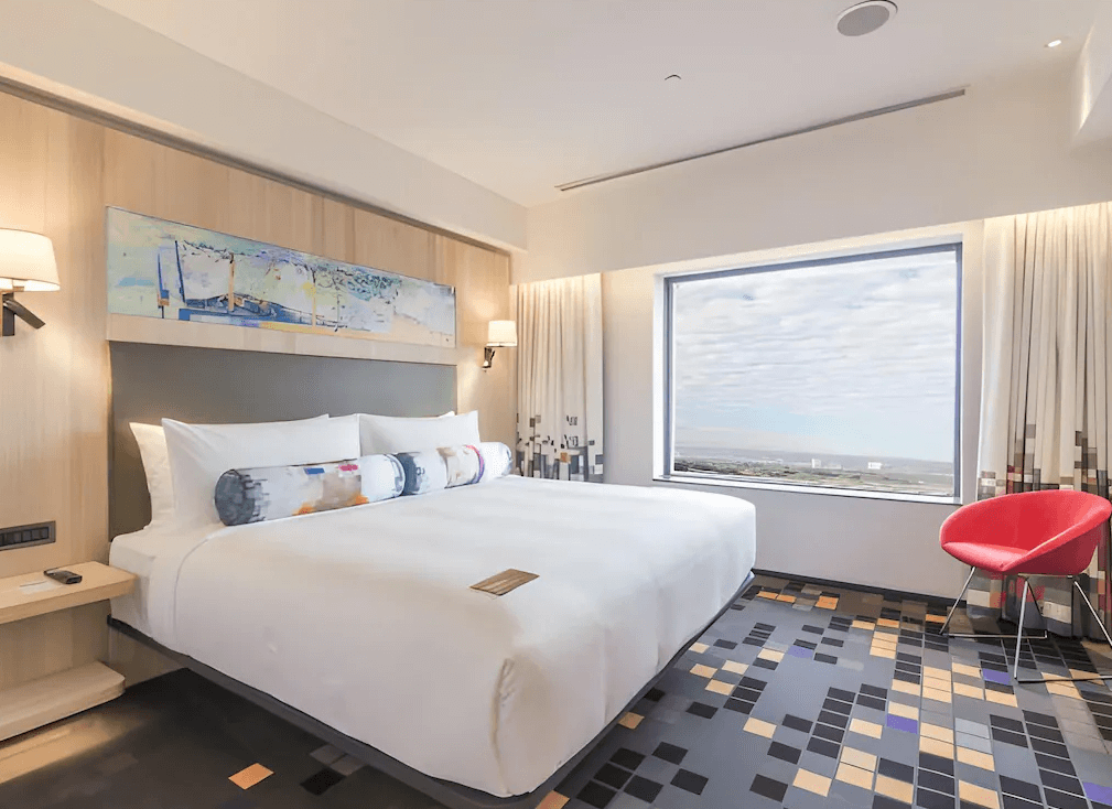 【台北寵物友善住宿】台北北投雅樂軒酒店的特大雙人床房型，房內光線明亮，搭配彩色抱枕與抽象畫作裝飾，窗外景致遼闊，配有紅色單椅與花磚地板，充滿現代活力氛圍。