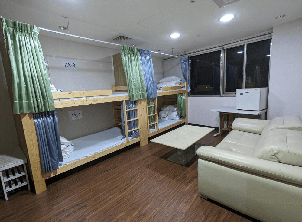 【三重住宿】台北橋黑熊好眠站膠囊旅館的多人房空間，配有上下舖床位，皆設有拉簾以保護個人隱私。房內還有皮沙發、茶几、小冰箱及對外窗，營造出舒適的共享住宿環境，適合背包客或團體旅客。
