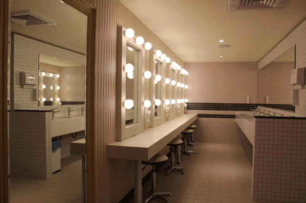 【三重住宿】台北橋黑熊好眠站膠囊旅館的公共梳妝空間，牆面以小方磚裝飾，設有多面鏡子與化妝燈，梳妝台前設有高腳椅。一旁為乾淨的洗手台區域，整體風格乾淨俐落，適合女性旅客使用。