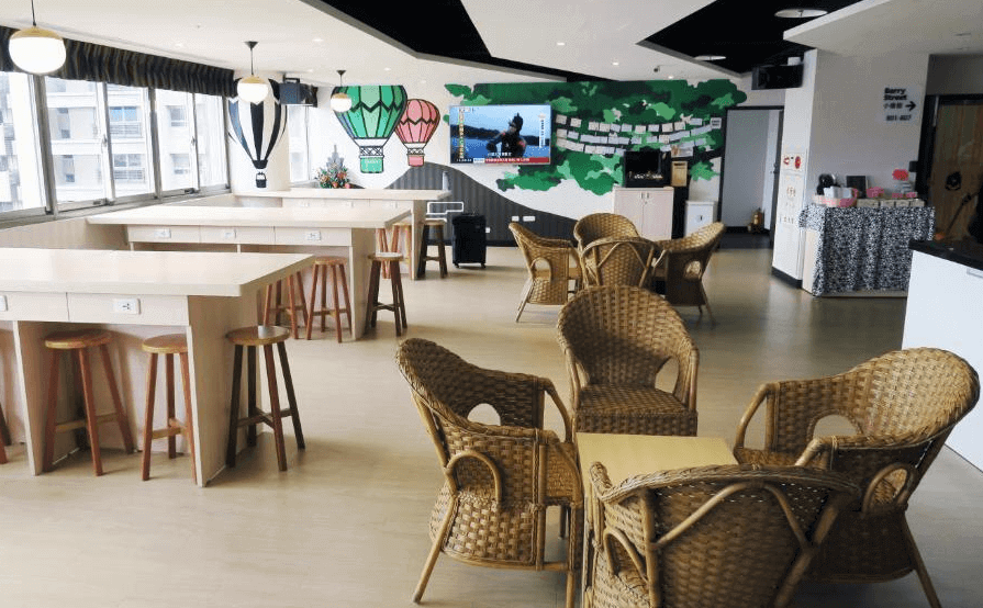 【三重住宿】台北橋黑熊好眠站膠囊旅館的交誼空間，配有藤椅組與高腳桌椅，牆面彩繪熱氣球與綠樹圖案增添趣味感。設有電視、插座與自然採光，適合用餐、交流或休息使用，氣氛輕鬆溫馨。