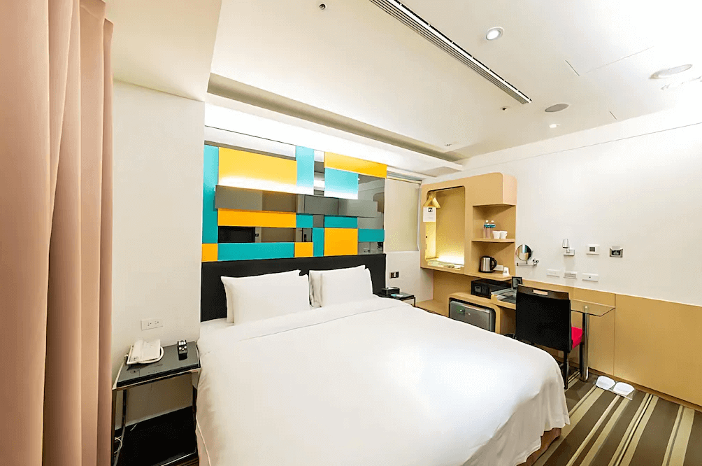 【蘆洲住宿】台北集賢商旅的雙人房，配有一張白色大床，床頭牆以彩色幾何設計增添活潑感。房內設有書桌、迷你冰箱、熱水壺及茶杯，整體空間明亮整潔，適合商務或休閒住宿。