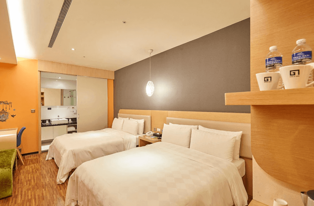 【蘆洲住宿】台北集賢商旅的家庭房配置兩張雙人床，牆面為深灰與橘色搭配，燈光柔和。房內備有書桌與開放式層架，後方可見簡易廚房空間，適合家庭或多位旅客入住。