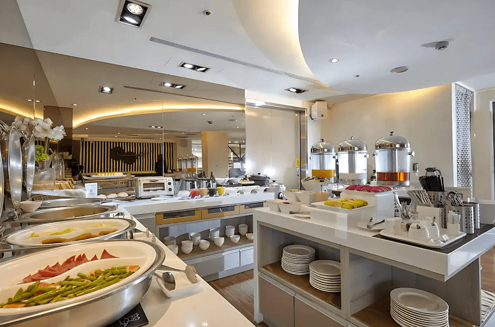 【蘆洲住宿】台北集賢商旅的自助早餐區，設有多種熱食、水果、果汁與咖啡機，餐檯整齊擺放白色瓷碗與餐具。整體空間明亮、動線流暢，提供旅客豐富多樣的早餐選擇。