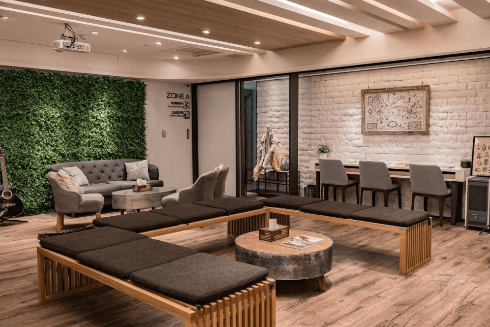 【台北車站青年旅館】台灣青旅膠囊旅店的交誼廳空間，設有灰色沙發、木質長椅與圓形茶几，牆面綠意植栽與裝飾畫點綴，另有書桌區與藝文擺設，適合旅客放鬆或社交交流。