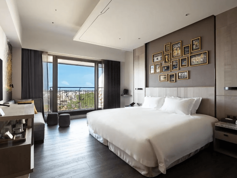 【龍潭住宿】名人堂花園大飯店的客房，配有一張加大雙人床，床頭牆面以多幅金框照片裝飾，提升藝術氣息。大片落地窗外即是城市與綠地美景，室內鋪設深色木地板，設有沙發、辦公桌與膠囊咖啡機，整體空間寬敞明亮，營造出舒適放鬆的住宿氛圍。