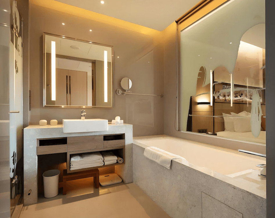 【龍潭住宿】名人堂花園大飯店的浴室空間，設有大型浴缸與鏡面洗手台，採用大理石與木質元素融合設計。鏡子兩側設有垂直燈條提供柔和照明，櫃體內整齊收納毛巾與體重計，浴缸旁的玻璃窗可俯瞰房內臥室區域，整體營造出高質感又舒適的泡澡體驗。