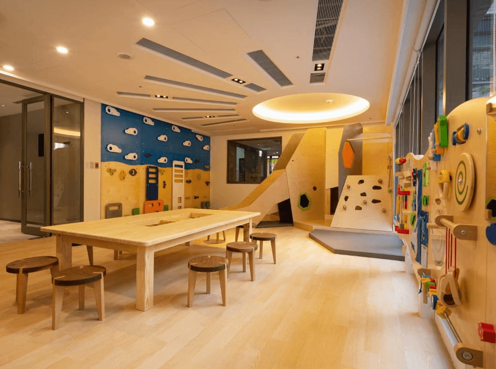 【龍潭住宿】名人堂花園大飯店的兒童遊戲室，設有原木質感的大桌子與小圓凳，牆面配有攀爬牆與各式互動遊戲設施，滑梯與攀岩牆設計兼具趣味與安全性。地板為淺色木質，空間整潔溫馨，適合親子同樂及小朋友安全活動。