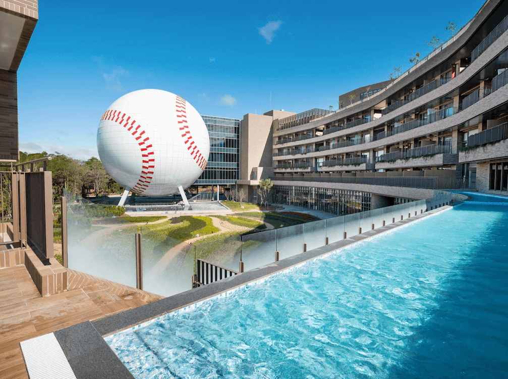 【龍潭住宿】名人堂花園大飯店的戶外景觀區域，設有造型弧形建築與透明玻璃圍欄的戶外泳池，水面清澈湛藍。遠處矗立一顆巨大白色棒球造型建築，象徵棒球主題特色，周邊綠意盎然，天空晴朗，整體景觀結合運動與休閒，極具特色與視覺吸引力。