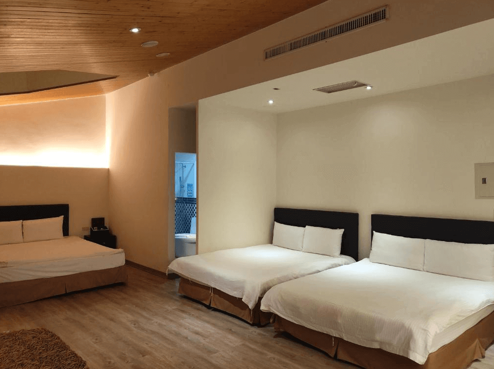 【楊梅住宿】君洋城堡的寬敞六人房空間，設有三張雙人床，天花板以木質造型延伸，搭配柔和燈光與淺米色牆面，營造溫馨舒適的住宿環境，浴室門外可見部分衛浴空間。