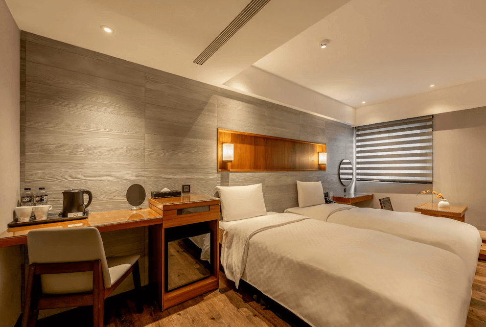【一中街住宿】宿野行旅的雙床房，配有兩張單人床，牆面以木紋灰色牆板設計，房內設有梳妝桌、電視與閱讀椅，窗邊亦設有榻榻米小茶几，氛圍舒適寧靜。