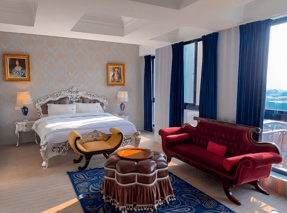 【草屯住宿】寶旺萊6號花園酒店的雙人房，銀白雕花床架搭配絨布紅沙發與歐式矮凳，牆上兩幅貴族畫作對稱擺設，藍色窗簾與地毯增添浪漫氣息，落地窗外可見陽台與遠方景致，整體空間明亮華麗。