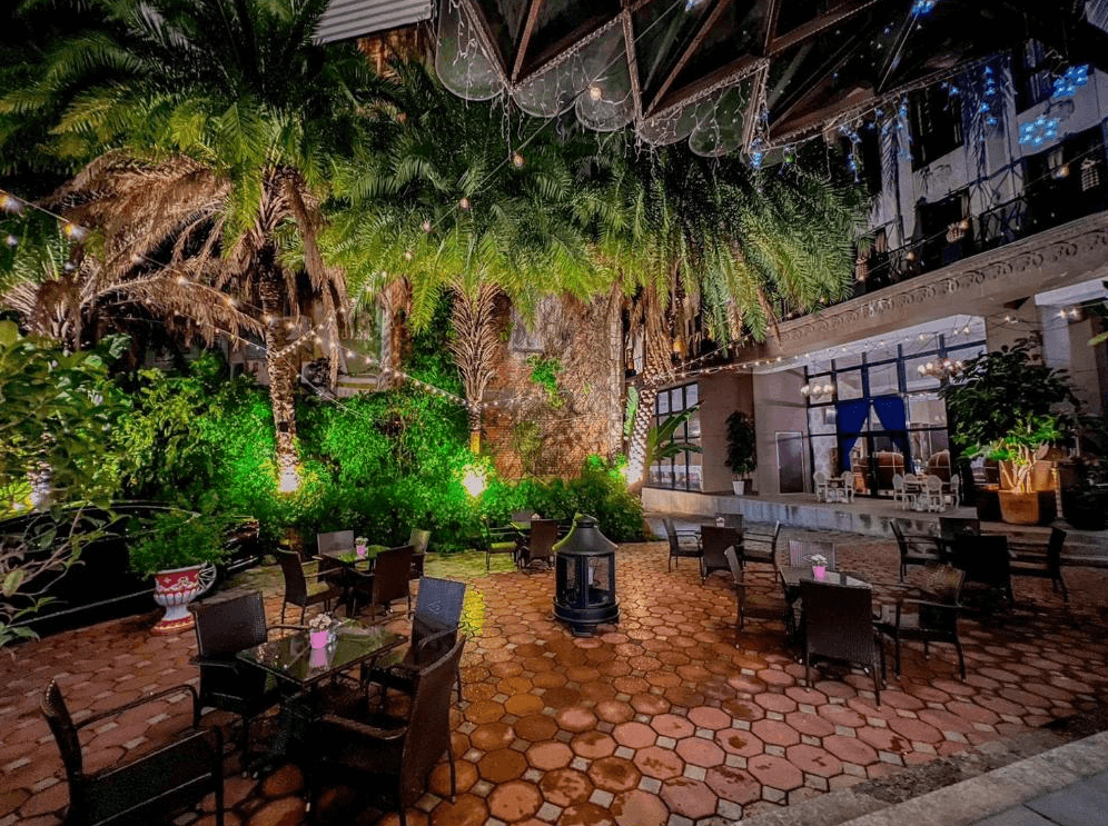 【草屯住宿】寶旺萊6號花園酒店的熱帶風格戶外花園，地面鋪設紅磚，四周環繞高大的棕櫚與綠植，設有多張玻璃桌椅可供用餐或喝茶，頭頂吊掛燈串增添浪漫氣氛，是放鬆談天的理想空間。
