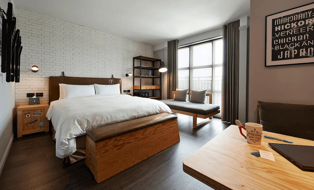 【三重住宿】捷絲旅 台北三重館的雙人房，採工業風裝潢，白色文化磚牆搭配深色木地板與皮革床頭。房內有一張雙人床、原木長椅、落地窗簾以及靠窗沙發區，整體風格現代簡約且舒適。