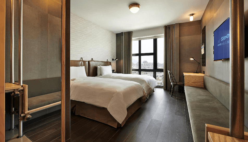 【三重住宿】捷絲旅 台北三重館，擁有兩張單人床的現代客房，空間寬敞，牆面為白磚與灰褐色面板混搭。設有大面積採光窗、木質書桌、工作燈與沙發座位，風格俐落實用，適合商務或好友同住。