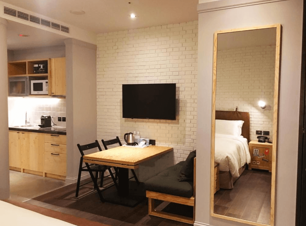 【三重住宿】捷絲旅 台北三重館的客房空間，配有簡易廚房（包含微波爐、電磁爐、咖啡機等），小型用餐區設有原木桌與黑色折疊椅。床區以白磚牆與溫暖燈光營造舒適感，適合長住旅客。