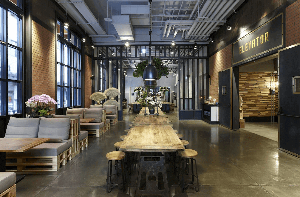 【三重住宿】捷絲旅 台北三重館的交誼廳，中央長桌搭配工業風高腳椅，兩側為木棧板沙發座位區，天花板裸露管線展現工業風格。空間寬敞、明亮，適合旅客休息或交流使用。