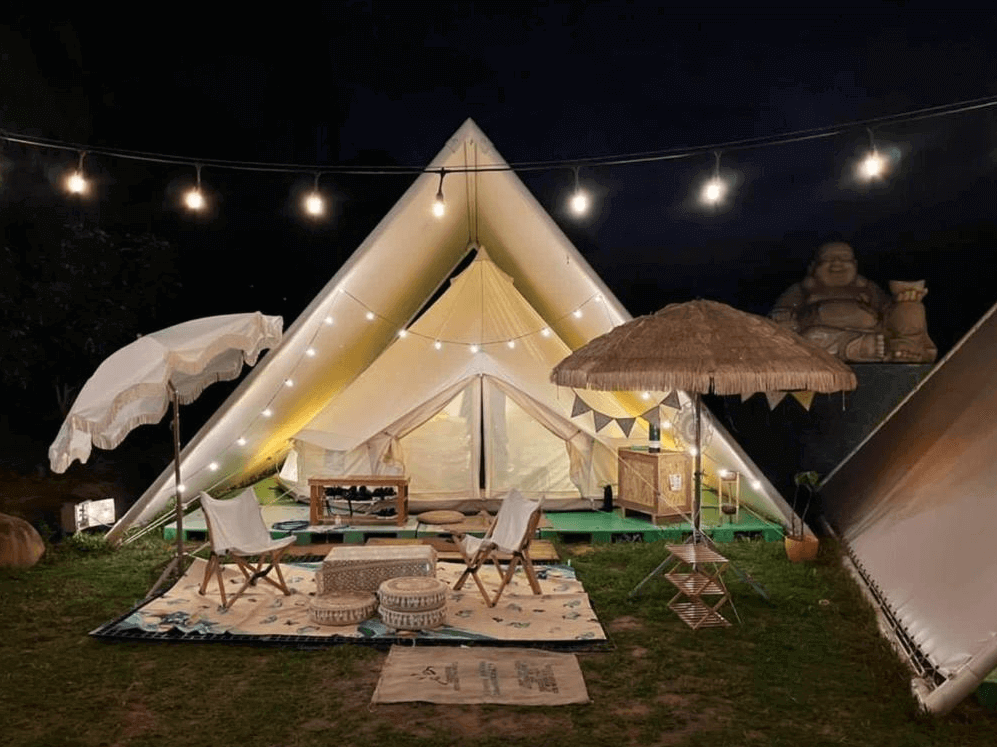 【龍潭住宿】晃晃印地安帳篷露營區的印地安帳篷在夜色中閃耀，帳篷前設有露天座位區與兩頂遮陽傘，地面鋪有地毯與坐墊，營造出慵懶悠閒的氛圍。上方掛滿串燈點綴整個空間，後方可見一尊大型笑佛雕像，增添露營區特色。