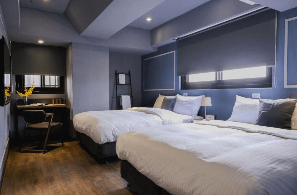 【三重住宿】柜富旅店的雙床房，以深藍色為主調，搭配木地板與柔和燈光。房內設有兩張舒適單人床，床邊窗戶設有遮光捲簾，牆角配置書桌與椅子，整體氛圍靜謐溫馨。