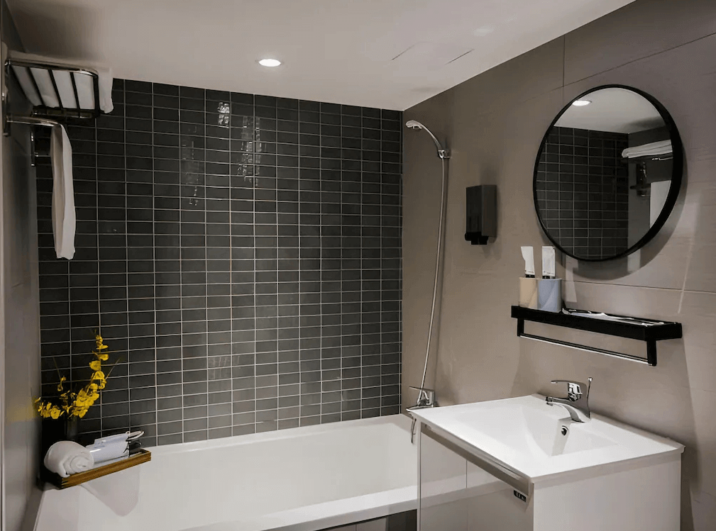 【三重住宿】柜富旅店的衛浴空間，設有白色浴缸與灰色磁磚牆面，圓形鏡面與簡約洗手台一體成型，並附上毛巾架與盥洗用品，整體設計簡潔實用。