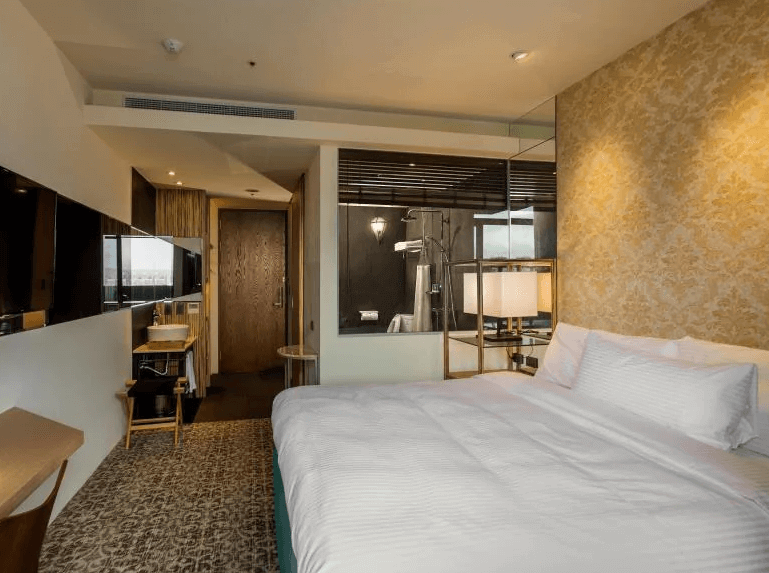 【蘆洲住宿】樂頤飯店的雙人房空間，設有一張白色床鋪與金色花紋牆面，床邊搭配玻璃隔間，內部可見乾濕分離的淋浴設備與現代燈飾。牆面掛有平面電視，整體風格典雅簡約。