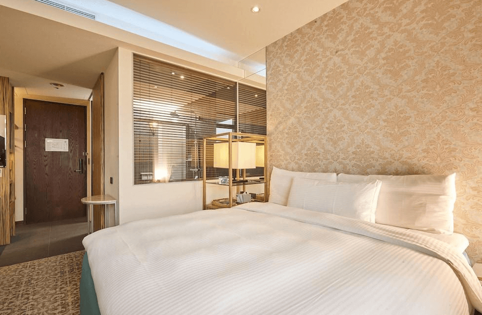 【蘆洲住宿】樂頤飯店的雙人房，白色床鋪與米金色壁紙營造溫馨感。床邊設有玻璃窗與百葉簾，可見後方乾濕分離的浴室區域。房內布置簡潔、舒適，光線充足。