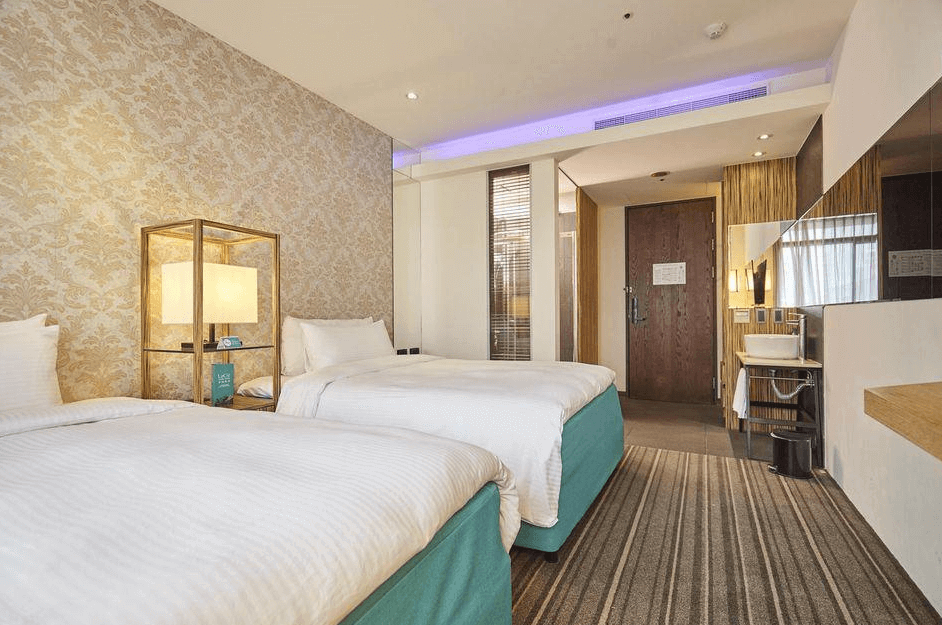【蘆洲住宿】樂頤飯店的雙床房，床鋪潔白、底座為綠松石色。牆面為典雅花紋設計，床邊設有黃銅色框架的現代風格立燈。空間寬敞，地毯設計為條紋與花紋混合風格，適合家庭或朋友同行入住。