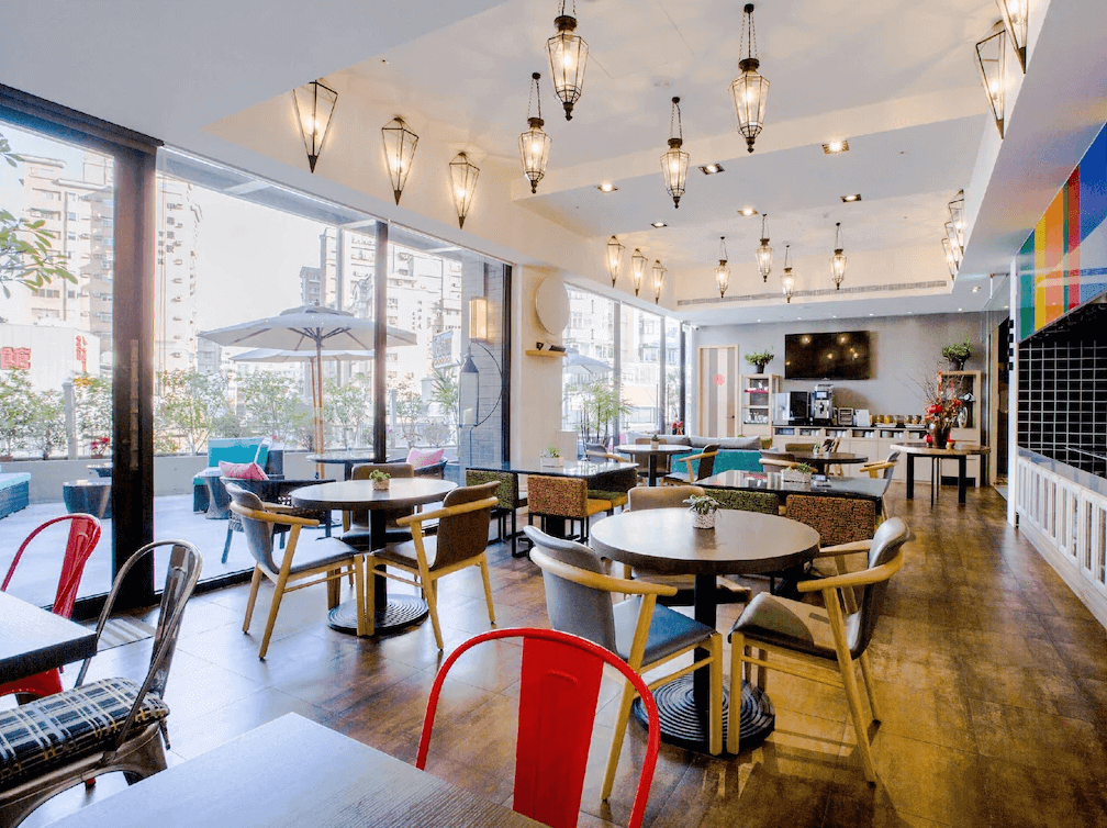 【蘆洲住宿】樂頤飯店的自助餐廳空間，落地窗引進大量自然光，室內裝潢明亮溫馨。設有多組圓桌與椅子，搭配各式風格坐墊與顏色。牆面配有咖啡機與早餐吧檯，整體氛圍輕鬆愜意，適合旅客用餐與放鬆。