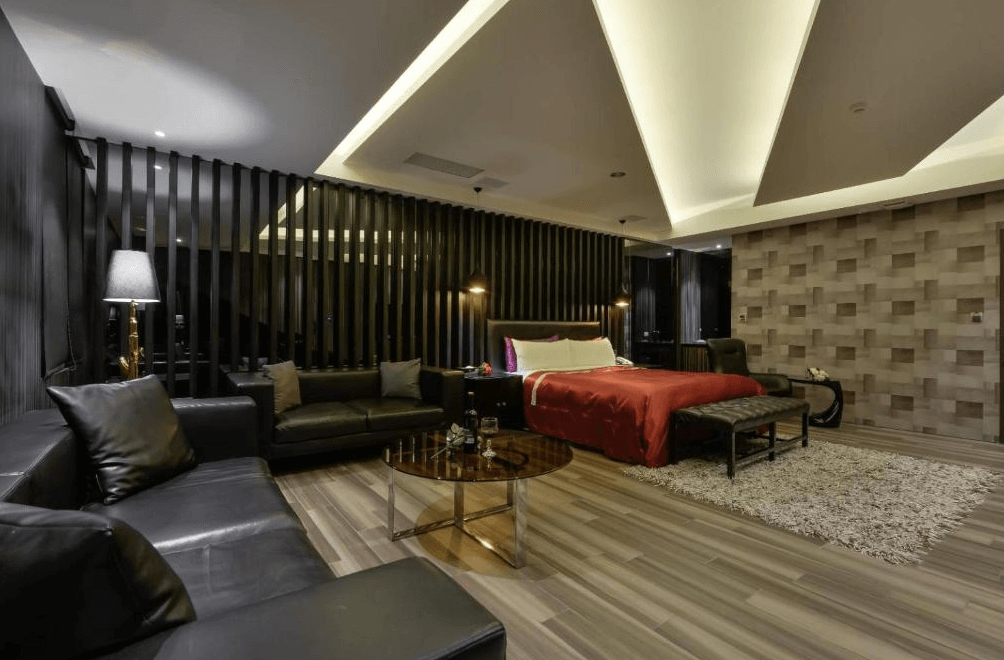 【草屯住宿】沐悅時尚精品旅館的現代豪華套房，房內配置一張大床與皮革沙發區，床鋪以紅色被鋪點綴，木質地板與天花板燈光設計營造層次感，牆面採用立體木紋與黑色格柵，時尚中帶有隱約奢華。