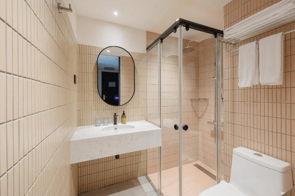 【台北寵物友善住宿】瀚寓夏天的衛浴空間，搭配淺米色磁磚與玻璃淋浴間，設有簡約白色洗手台、圓形鏡與置物層架，空間明亮清潔，設計俐落實用。