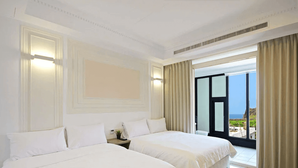 【綠島住宿】綠島House16的雙床房，牆面裝飾立體線條設計，整體以白色調為主，搭配柔和燈光與大片窗簾，窗外可遠眺海景與山巒。