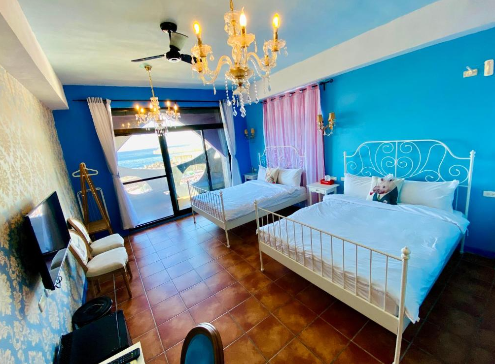 【綠島寵物友善住宿】綠島寧靜海潛水-城堡館藍白相間的地中海風格四人房，兩張白色鐵藝雙人床搭配水晶吊燈與復古壁紙，大片落地窗外可遠眺海景，室內明亮清新，充滿渡假氣息。