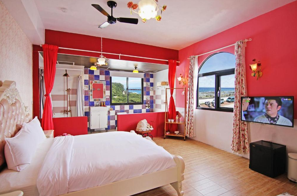 【綠島寵物友善住宿】綠島寧靜海潛水-城堡館，以紅色與粉色為主調的浪漫雙人房，設有雕花床架與木質地板，浴室區採開放式設計，搭配彩色磁磚牆與大片窗戶，可眺望窗外海景，營造出溫馨舒適氛圍。