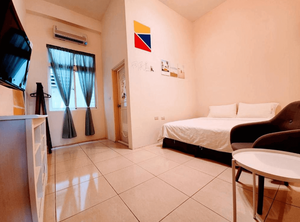 【綠島住宿】綠島彩虹民宿的雙人房，牆上有幾何拼貼與簡約掛畫，設有雙人床、沙發椅與圓桌，落地窗透入自然光線，適合情侶或單人旅客。