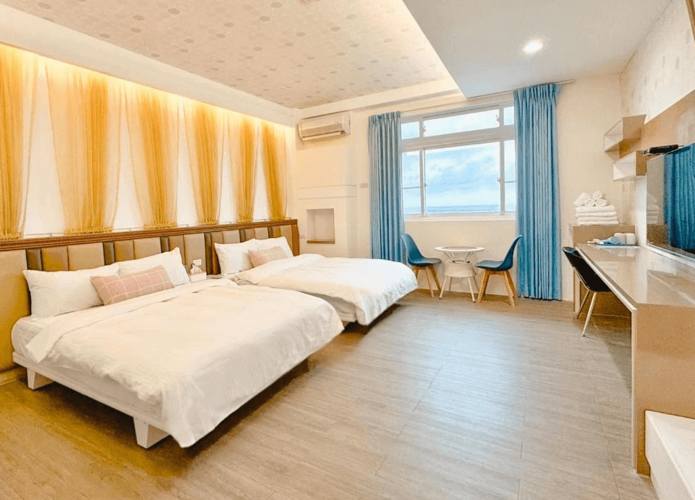 【綠島住宿】綠島海灣會館的四人房，設有兩張大床與柔和燈光牆面，搭配大片海景窗與藍色窗簾，自然光灑落房內，展現舒適放鬆的氛圍。
