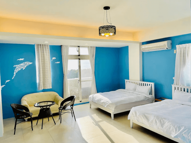 【綠島住宿】綠島藍漾海墅的四人房，藍白海洋風設計風格，搭配兩張白色雙人床、藤編椅與沙發，牆面有海豚壁貼裝飾，營造出清新愜意的度假氛圍。