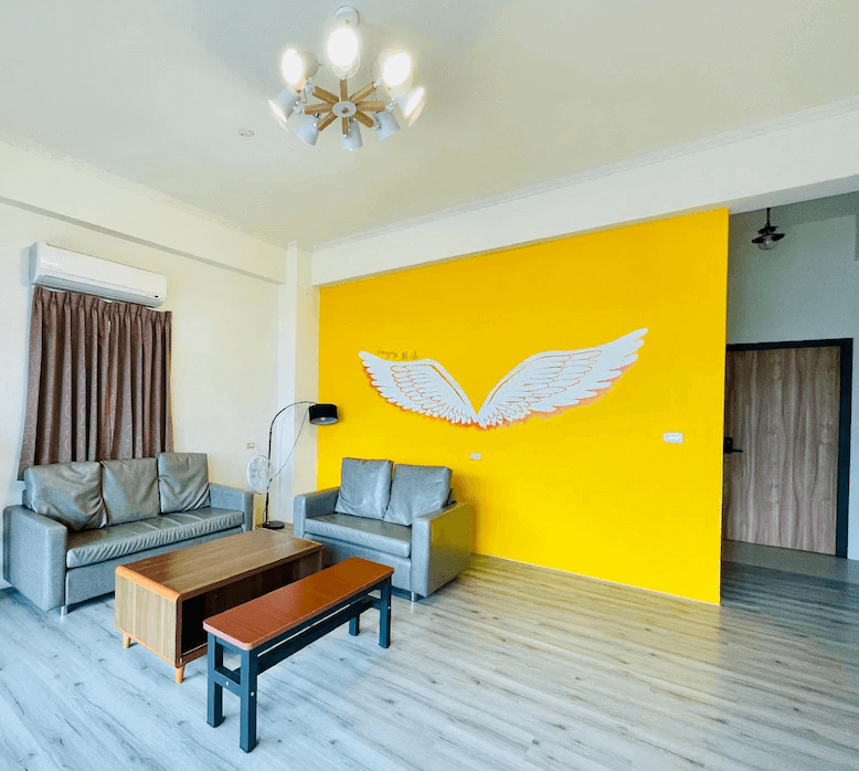 【綠島住宿】綠島藍漾海墅的客廳空間，亮黃色牆面上裝飾一對白色天使翅膀圖樣，搭配灰色沙發與原木茶几，木質地板營造溫馨且活潑的社交空間。