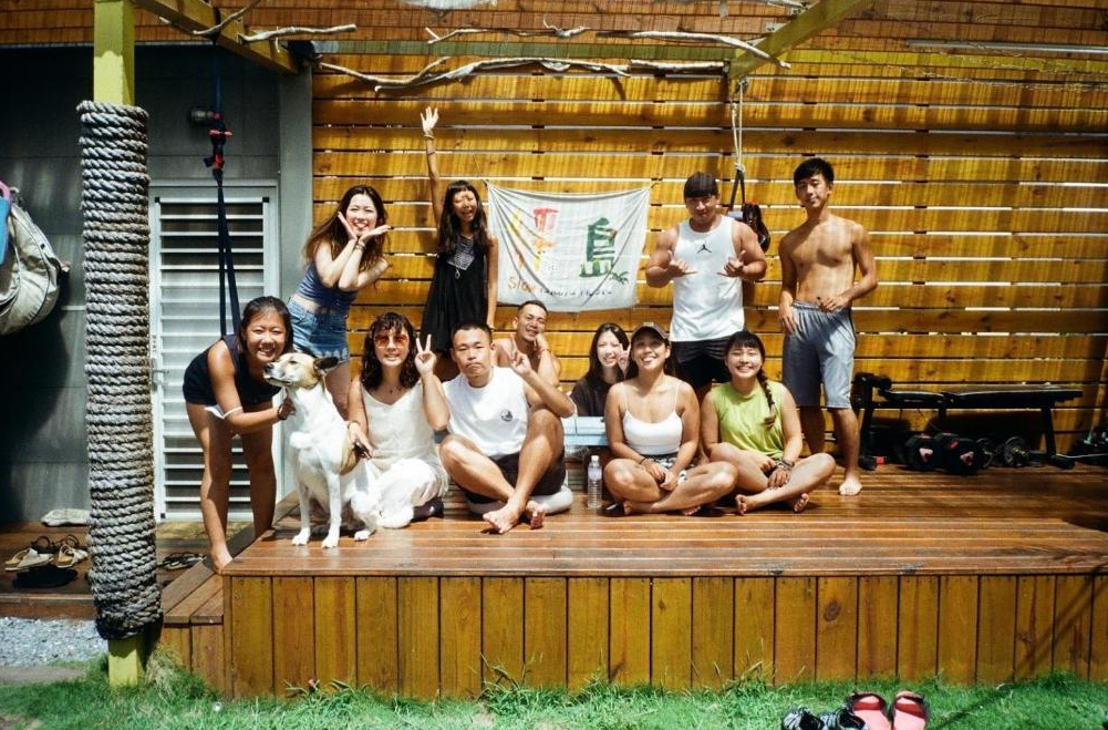 【綠島寵物友善住宿】"綠島住宿"緩島旅宿，一群開心的年輕旅人與一隻狗狗，在木質平台上合影，背景為旅宿旗幟與健身器材，展現友善熱情與社群氛圍。