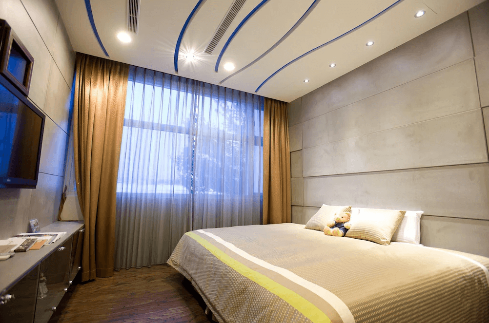 【草屯住宿】草屯知達工藝會館的雙人房，牆面以米灰色系搭配質感木地板，天花板裝飾有藍色弧線燈帶，床鋪擺放整齊，床上放有可愛泰迪熊娃娃，大片窗簾採光良好，整體空間明亮溫馨。