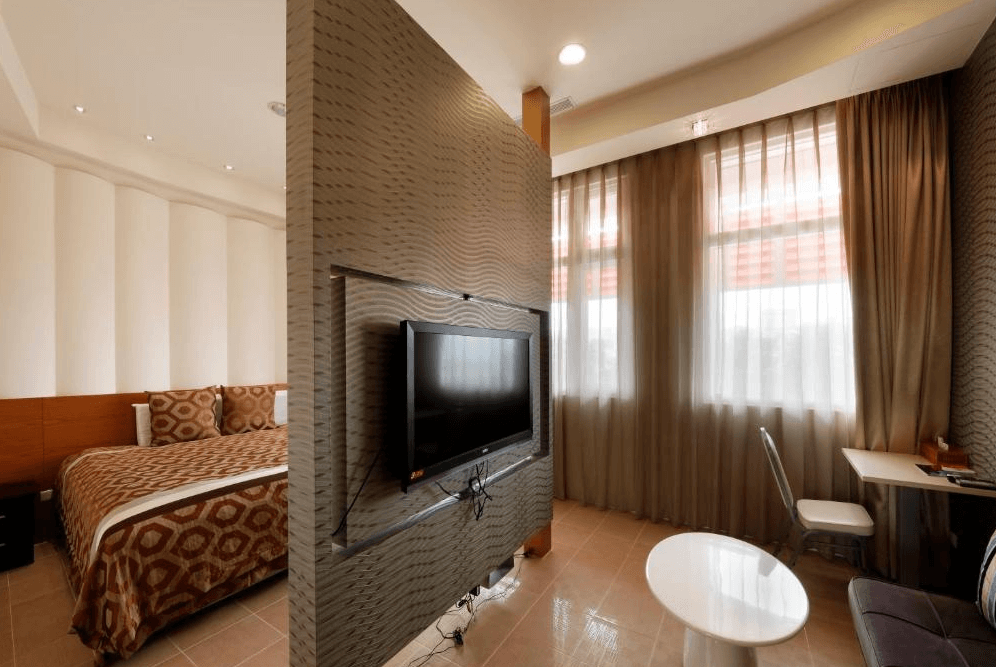 【草屯住宿】草屯知達工藝會館的臥房與客廳分隔，床鋪區鋪有幾何圖案床罩，客廳區配有桌椅與電視，大片窗戶搭配咖啡色窗簾，室內採光佳，空間動線清晰、適合簡單辦公或休憩。